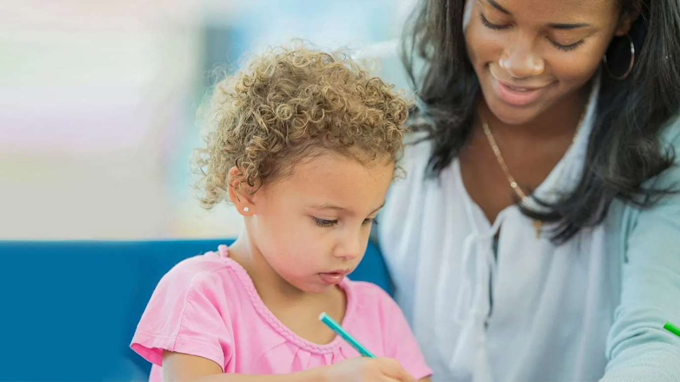 An African American woman helps a young girl use colored pencils.