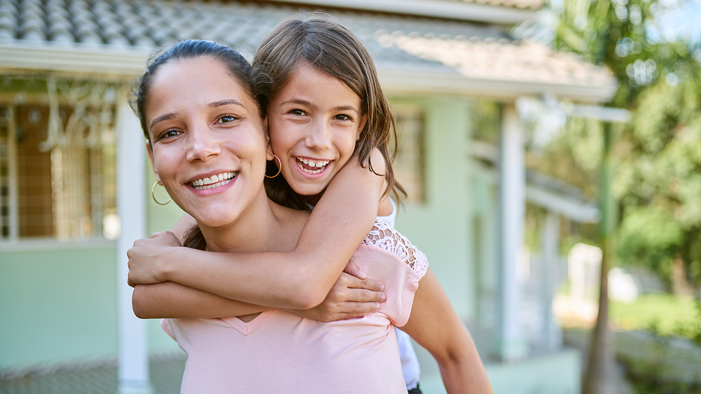 A BIPOC woman gives her daughter a piggy back ride in front of a small green house.