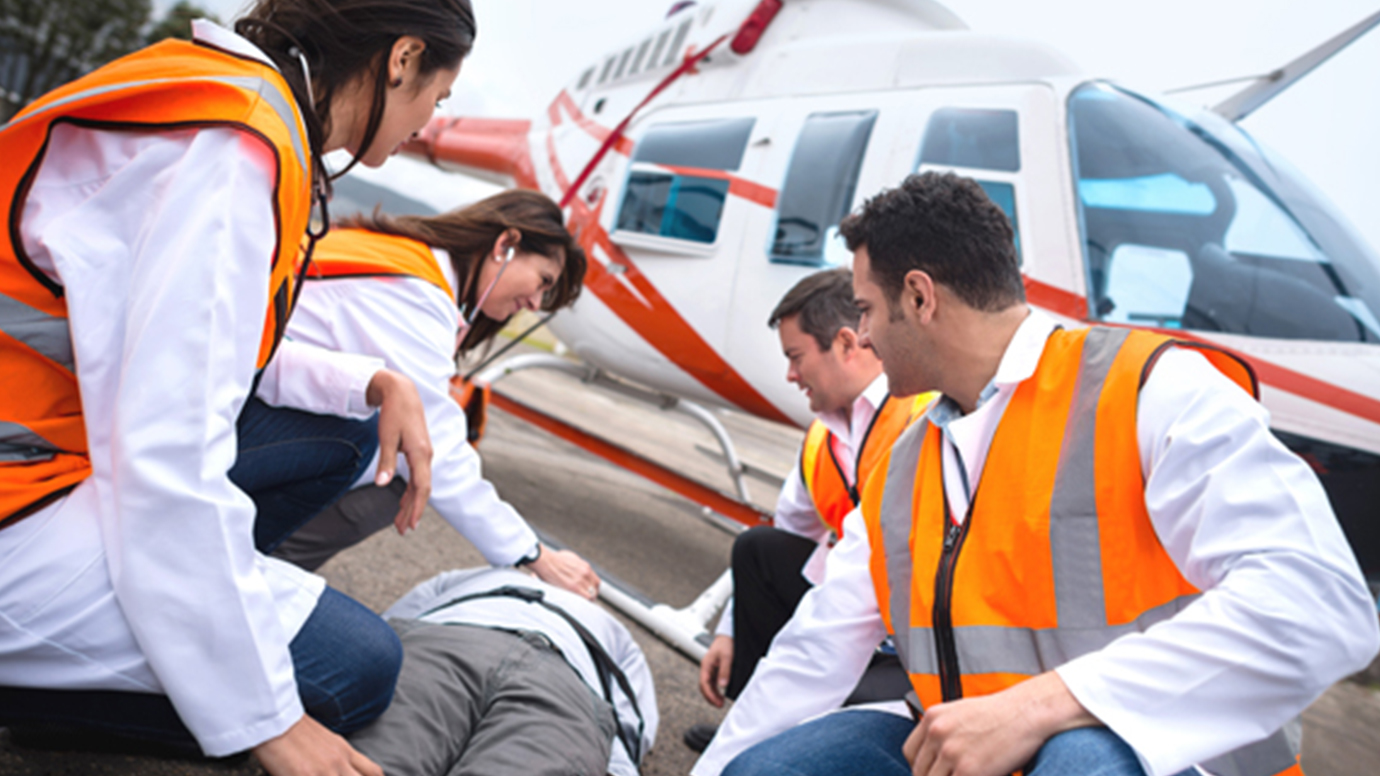 Four first responders in safety gear surround a man lying on a stretcher with a medical helicopter in the background.